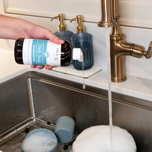 A hand holds a bottle of PROV 31 Dish Soap over a kitchen sink with running water, soap bubbles, and two blue soap dispensers on the counter.
