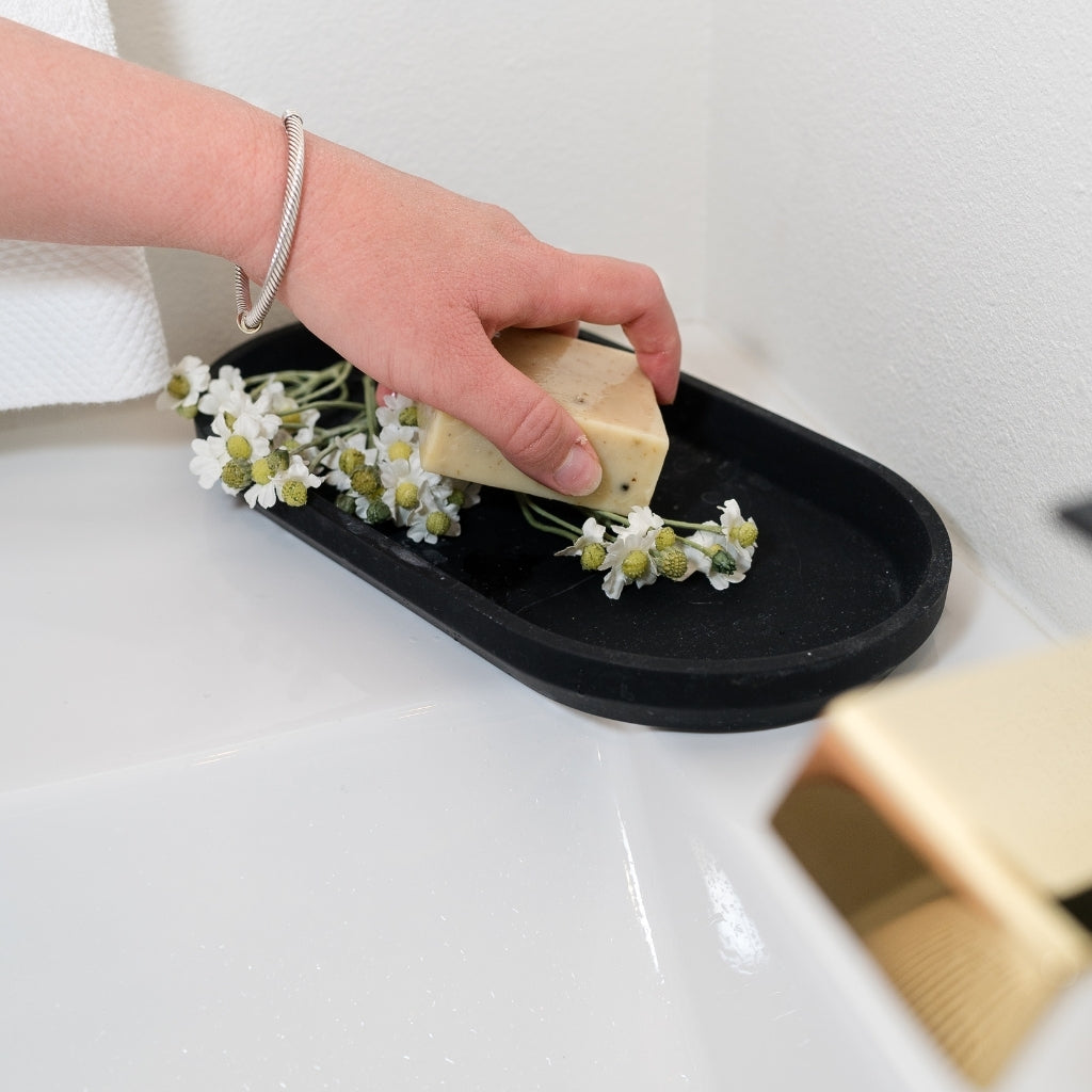 A hand holding a bar of PROV 31 fragrance free soap over a black oval tray with chamomile, placed on a white sink. A gold soap dish is partially visible in the foreground.
