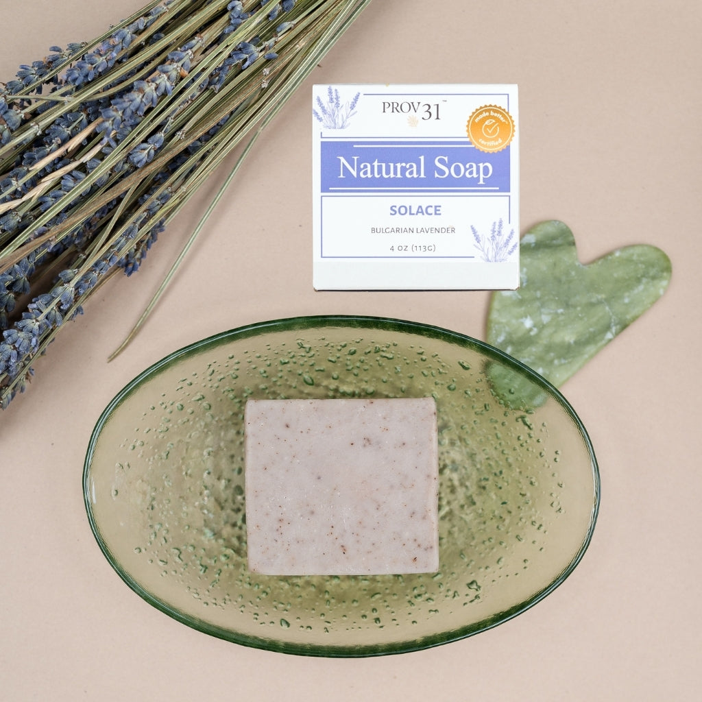 A square bar of PROV 31 Natural Soap labeled 'Solace' sits in a translucent green oval dish, styled with dried lavender stems and a green heart-shaped stone on a beige background.