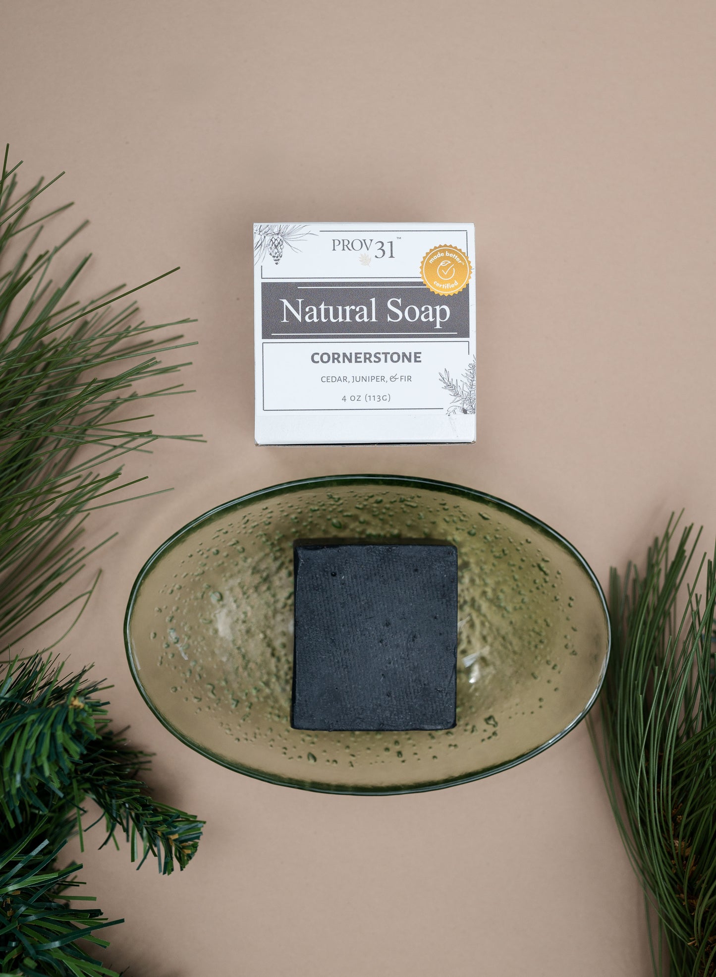 A bar of PROV 31 soap in a speckled green bowl with branded box packaging above, styled with pine branches on a beige background. The box reads 'PROV 31 Natural Soap Cornerstone'.