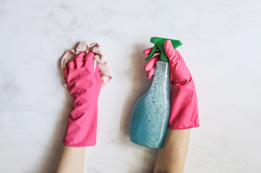 Person wearing pink gloves holding a blue cleaning spray bottle on a light background