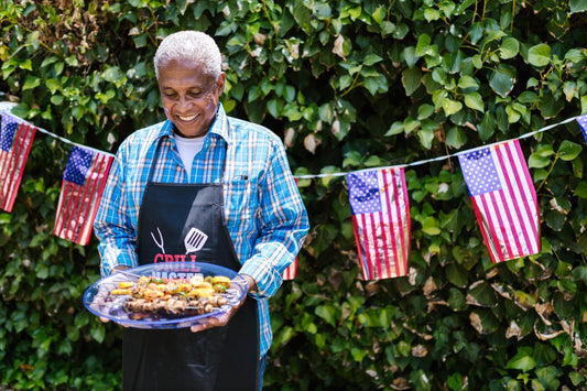 Man holding a plate of grilled food in front of a green hedge with American flags.