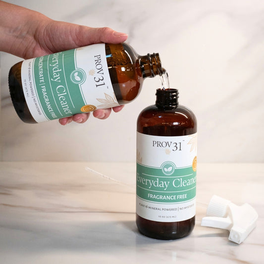 A hand pours liquid from an amber bottle labeled PROV31 Everyday Cleaner into a second amber bottle with a white spray nozzle cap placed beside it on a light marble surface.