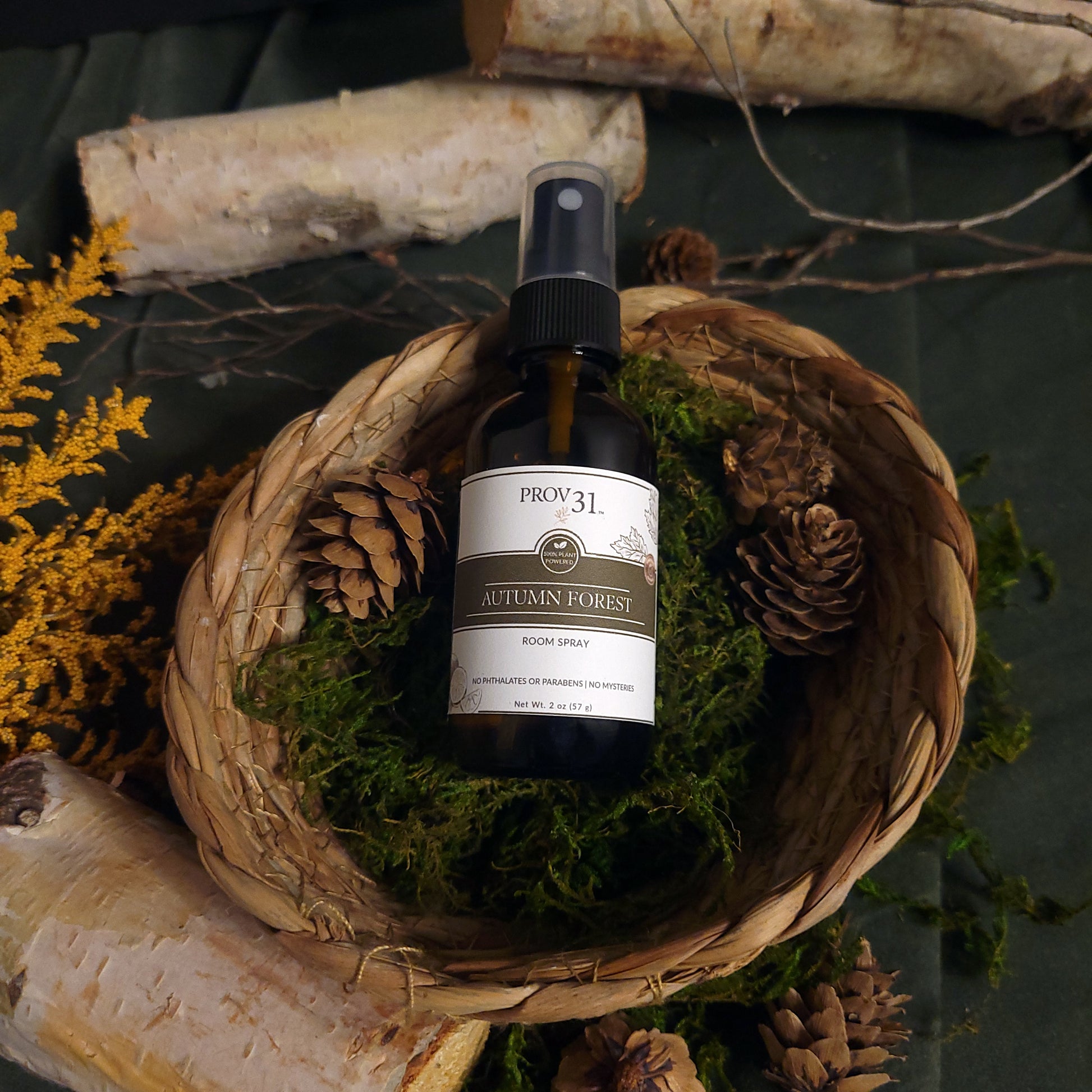 Bottle of Prov 31 Autumn Forest room spray in a woven basket with moss and pine cones.