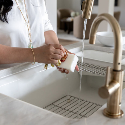 A woman in a white blouse scrubs a dish cloth with PROV 31 Dish Soap Block under running water from a tall brushed-gold faucet in a modern kitchen sink, next to clean white dishes.