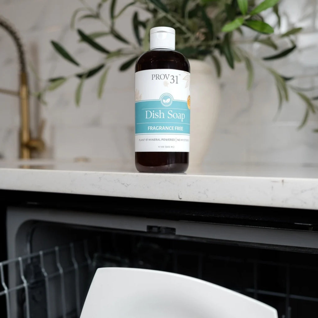 A bottle of Prov 31 Dish Soap, fragrance-free, standing on a white marble kitchen countertop with a dishwasher below and a potted plant in the background.