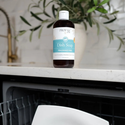 A bottle of Prov 31 Dish Soap, fragrance-free, standing on a white marble kitchen countertop with a dishwasher below and a potted plant in the background.