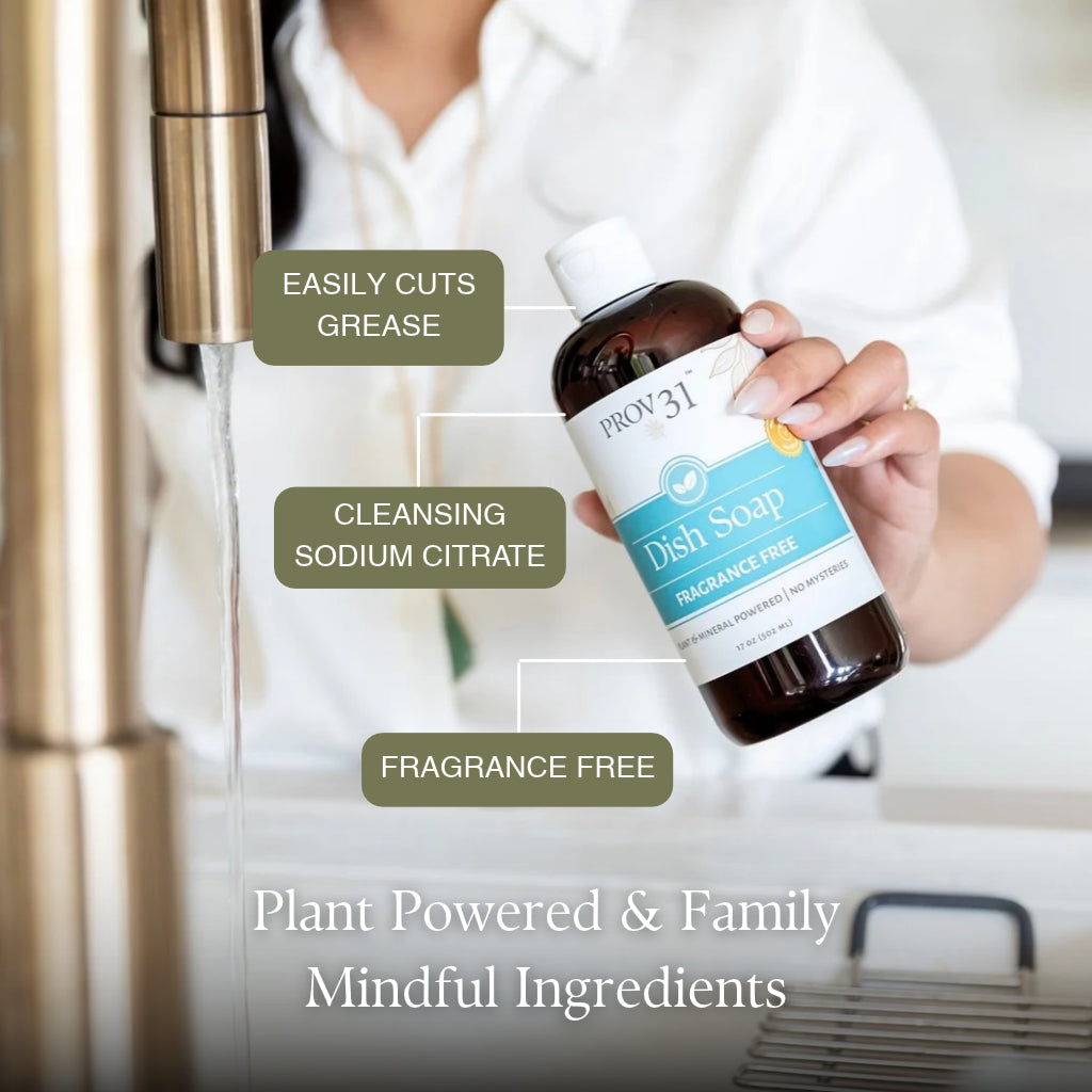 A person in a white shirt holds a bottle of Prov31 Dish Soap under a running faucet in a kitchen setting. The bottle is dark amber with a white cap and a label that is light blue and white. Text callouts read easily cuts grease, cleansing sodium citrate, and fragrance free.