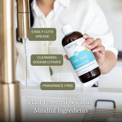 A person in a white shirt holds a bottle of Prov31 Dish Soap under a running faucet in a kitchen setting. The bottle is dark amber with a white cap and a label that is light blue and white. Text callouts read easily cuts grease, cleansing sodium citrate, and fragrance free.