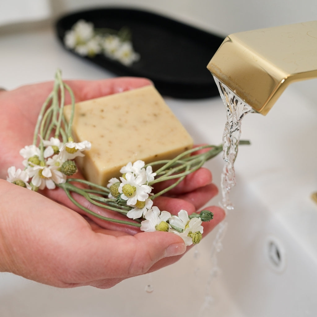 A hand holds a rectangular bar of pale beige bar soap decorated with chamomile flowers under running water from a gold faucet in a white sink.