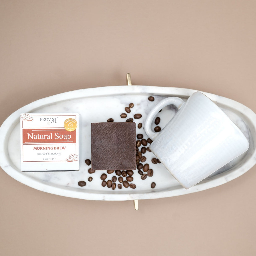 A styled flat-lay of PROV 31 bar soap and its packaging on a white marble tray, next to a white ceramic mug and scattered coffee beans, on a solid beige background. 