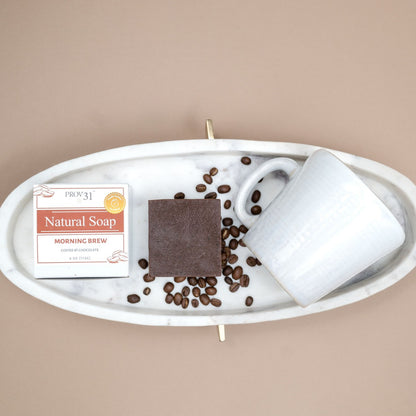 A styled flat-lay of PROV 31 bar soap and its packaging on a white marble tray, next to a white ceramic mug and scattered coffee beans, on a solid beige background. 