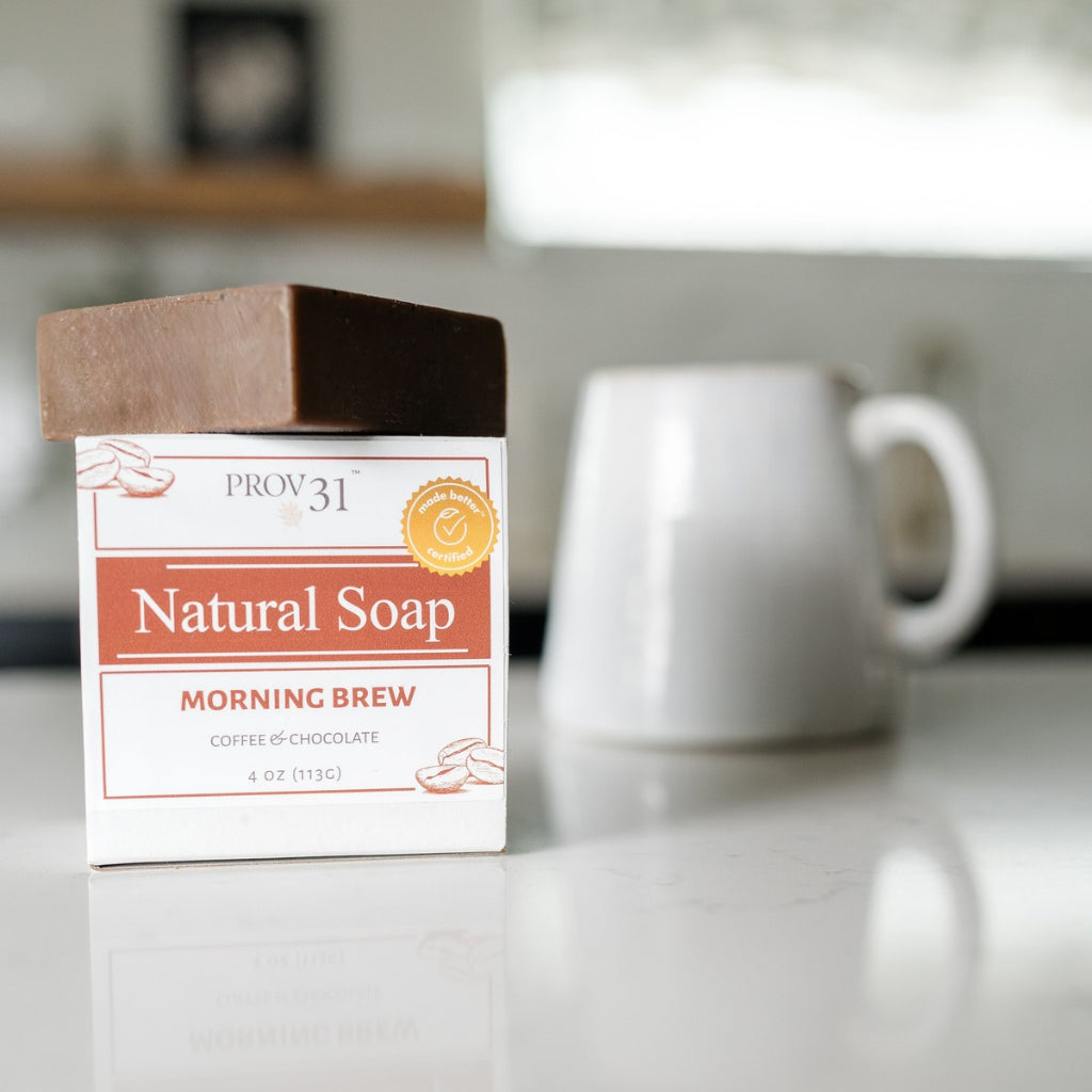 A bar of PROV 31 Natural Soap called Morning Brew sits on a white kitchen countertop with a blurred white ceramic mug in the background. The soap bar sits on top of its white box packaging.