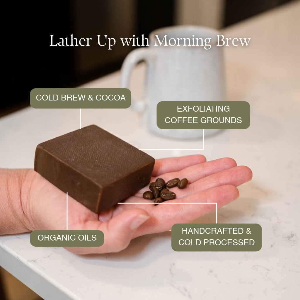 A hand holds a rectangular bar of dark brown PROV 31 soap with coffee beans on the palm, set against a kitchen counter with a mug in the background. Text callouts describe the soap as cold brew and cocoa, exfoliating coffee grounds, with organic oils and handcrafted cold processing.