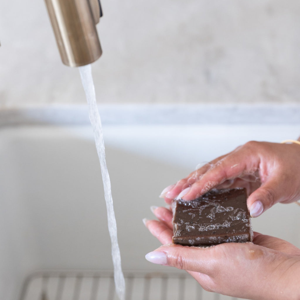 A close-up of a woman's hands washing a dark brown rectangular bar of PROV 31 natural soap under running water from a brushed brass faucet in a sink.