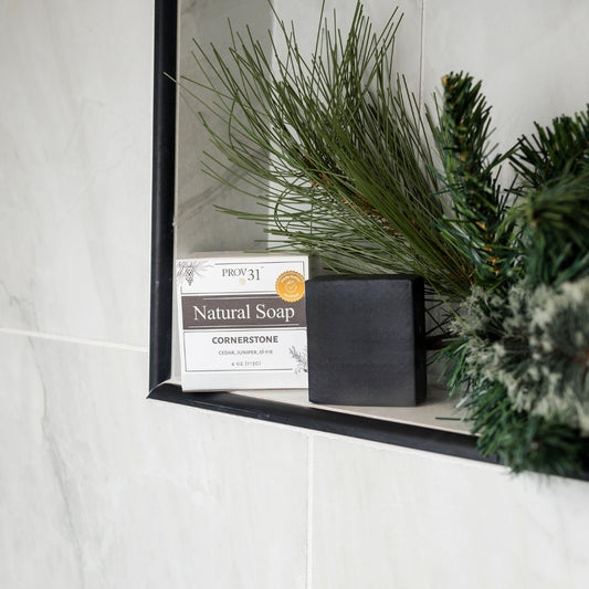 A box of PROV 31 Natural Bar Soap in the Cornerstone scent sits next to a black charcoal soap bar on a shower shelf, styled with pine branches against a white marble wall.