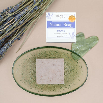A square bar of PROV 31 Natural Soap labeled 'Solace' sits in a translucent green oval dish, styled with dried lavender stems and a green heart-shaped stone on a beige background.