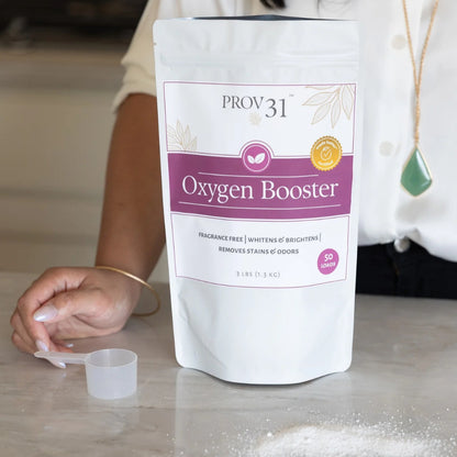 A stand-up pouch of Prov 31 Oxygen Booster placed on a kitchen counter, with a woman's hand and torso visible behind it. The package is white with a magenta label and a small clear scoop and the powder in front.