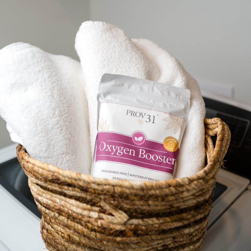A bag of PROV 31 Oxygen Booster placed inside a woven basket with white towels, set against a softly blurred indoor background of a washing machine.
