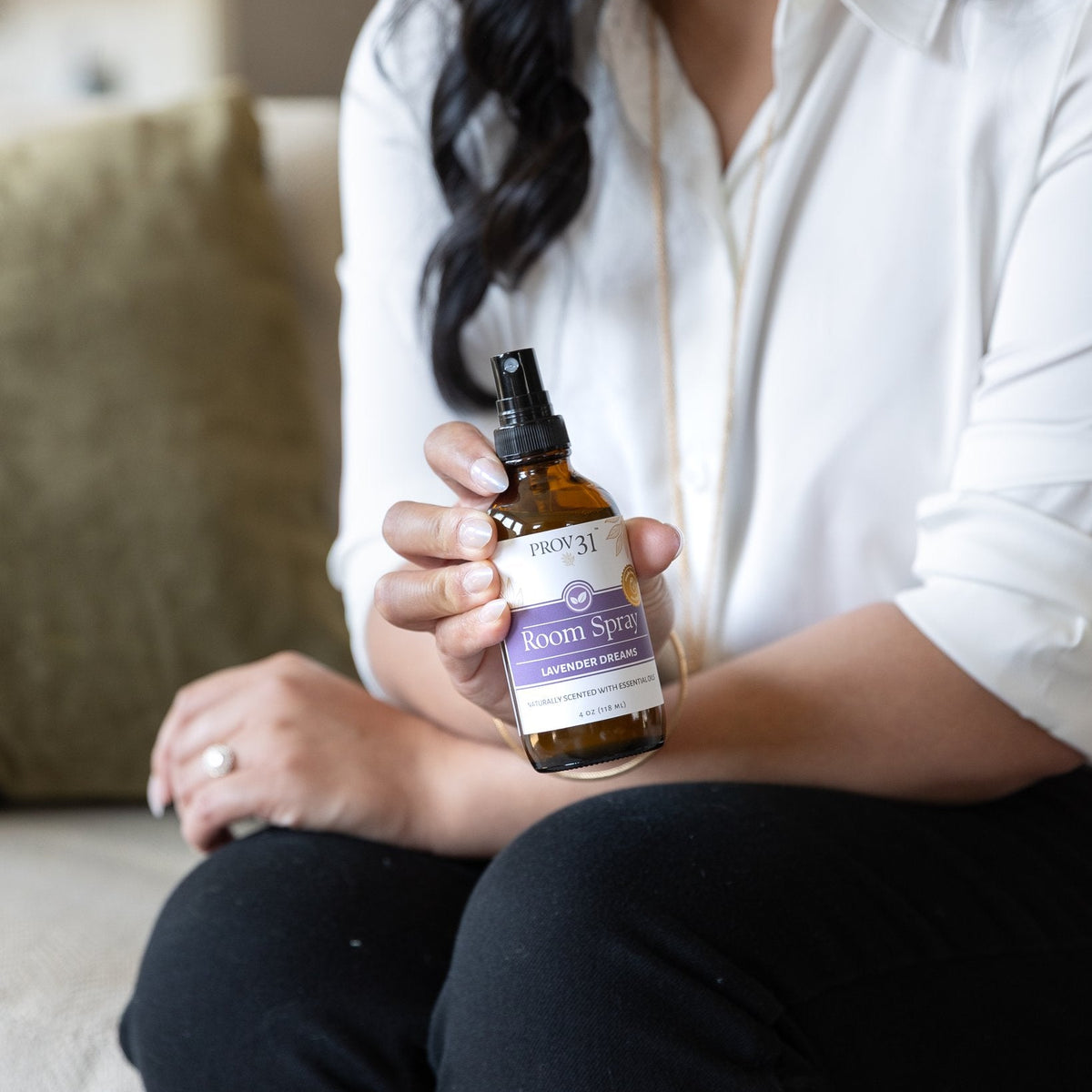 A woman in a white button-down shirt and black pants holds a small amber glass PROV 31 lavender room spray bottle with a black spray top and a purple-and-white label. The setting appears to be a softly lit indoor room with a cushion in the background.