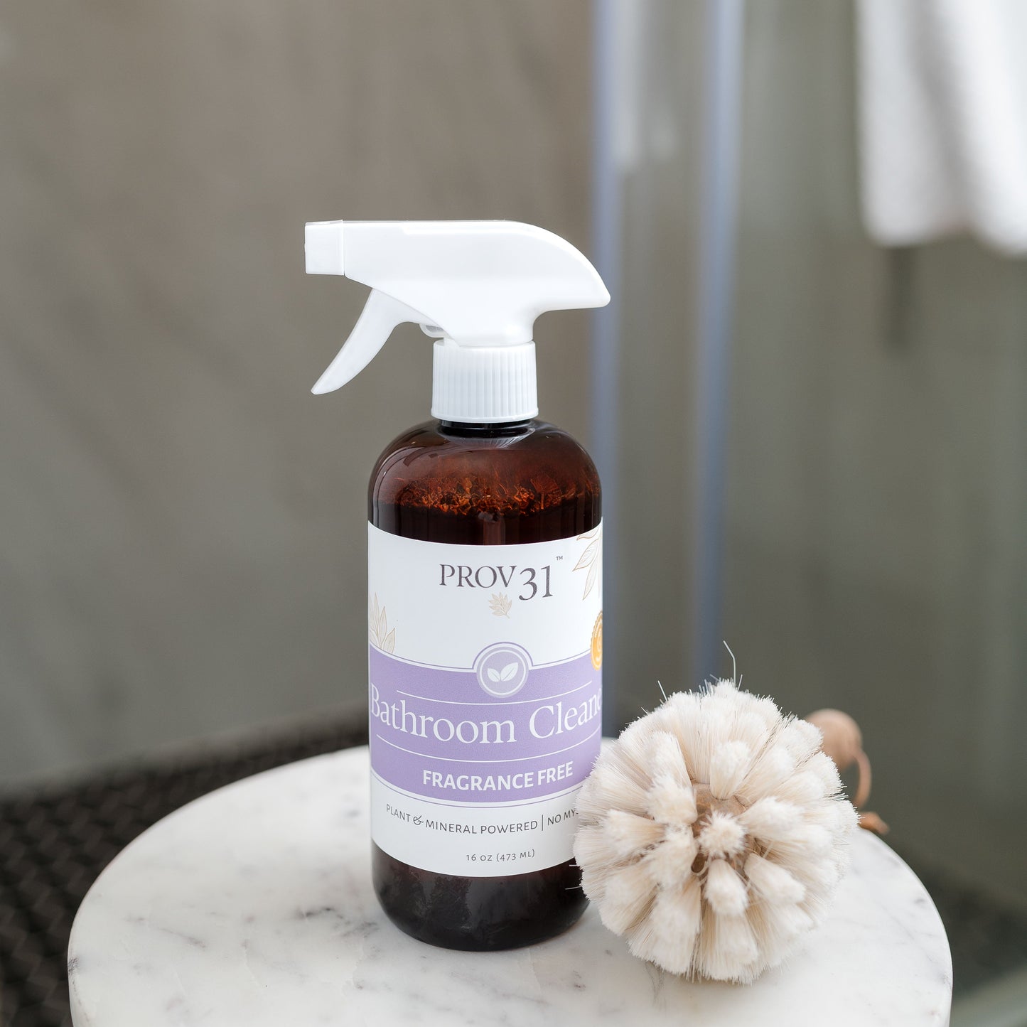 A bottle of PROV 31 Bathroom Cleaner, fragrance-free, with a white spray trigger top and a purple label, sitting on a white marble table next to a wood scrub brush in front of a shower.