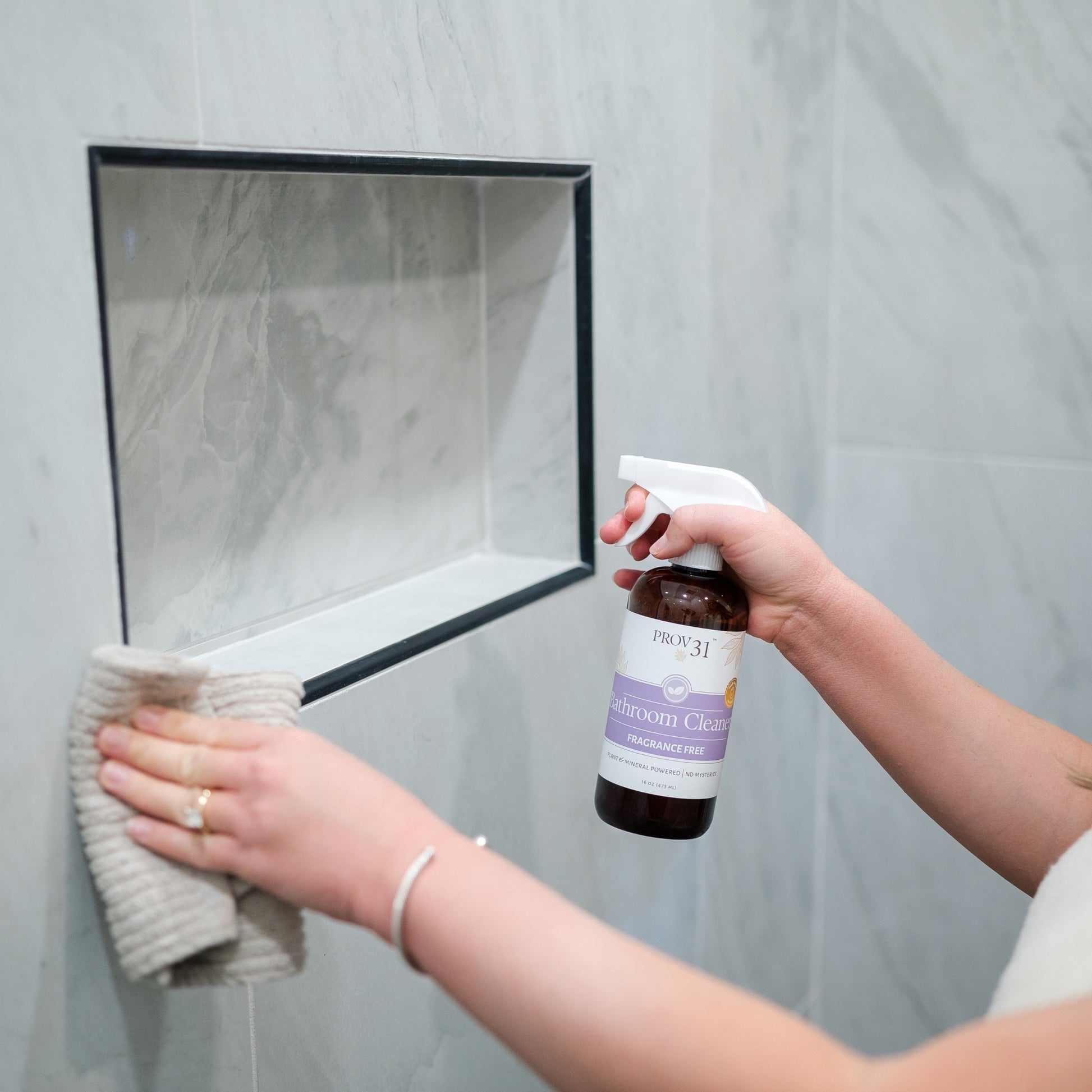 A person in a bathroom holds a bottle of PROV 31 bathroom cleaner spray while wiping a gray marble-tiled wall with a beige cloth.