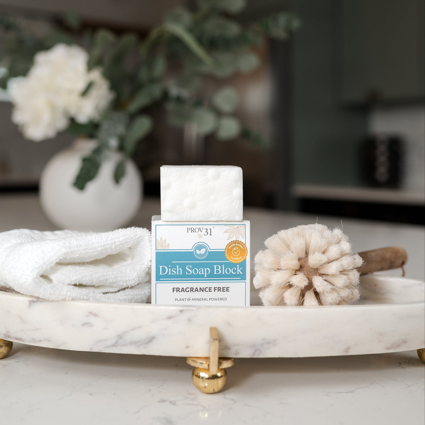 PROV 31 dish soap block sits on a white marble tray with gold feet, styled with a folded white towel and a wooden bottle brush in a bright kitchen setting.