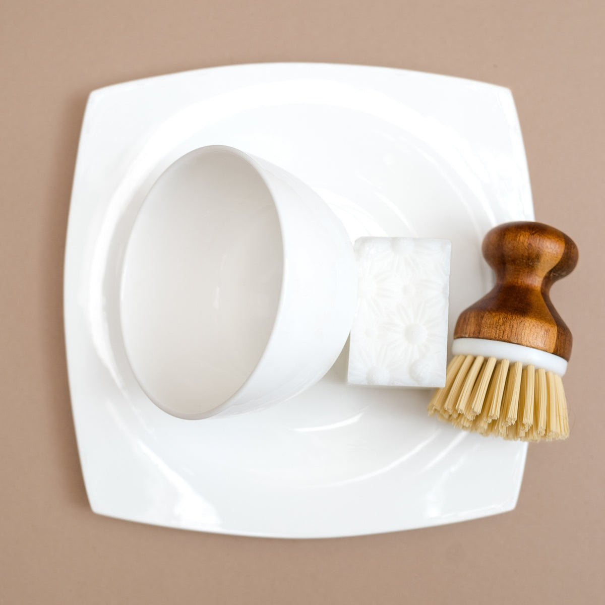 A white ceramic bowl with a matching square plate, a white textured soap bar, and a wooden-handled brush with natural bristles, arranged on a plain beige background.