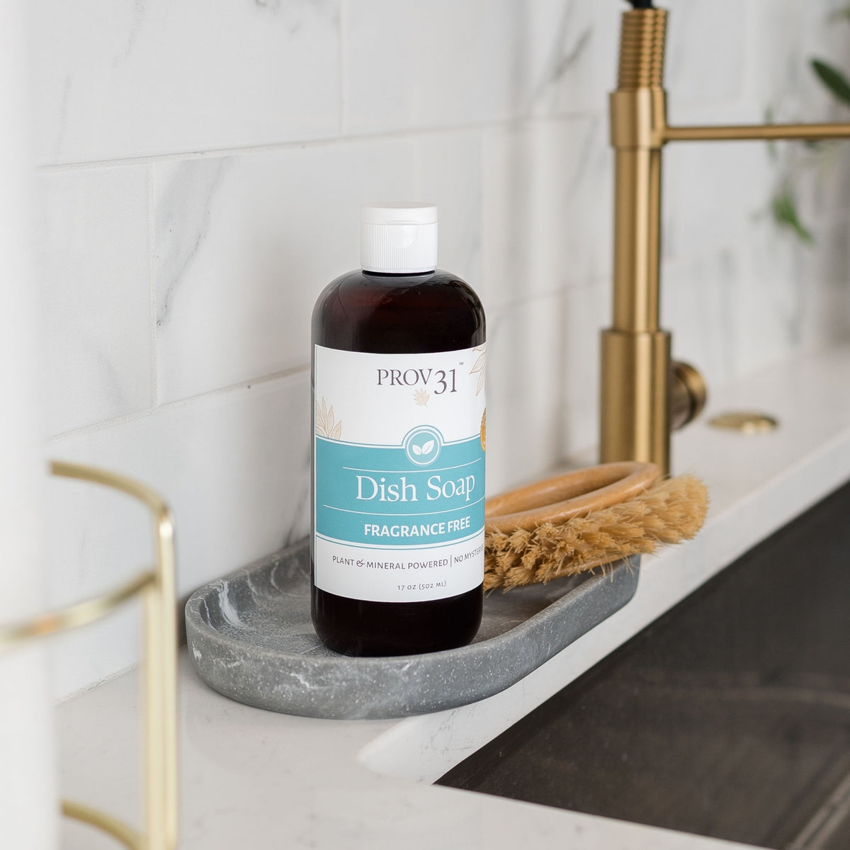 A bottle of Prov31 Dish Soap, fragrance-free, standing on a gray marble dish rack next to a wooden scrubber in a modern kitchen sink with a gold faucet.