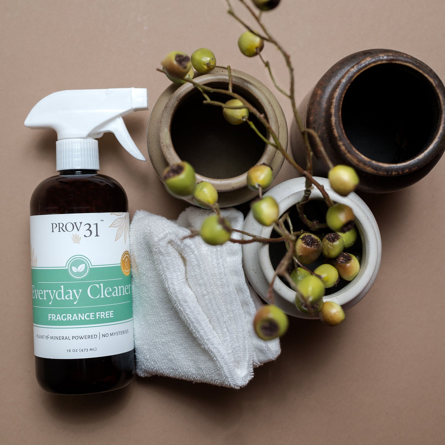 A bottle of PROV 31 Everyday Cleaner spray with a white trigger sprayer and a white cloth, styled with ceramic pots and green berry branches on a solid beige background.