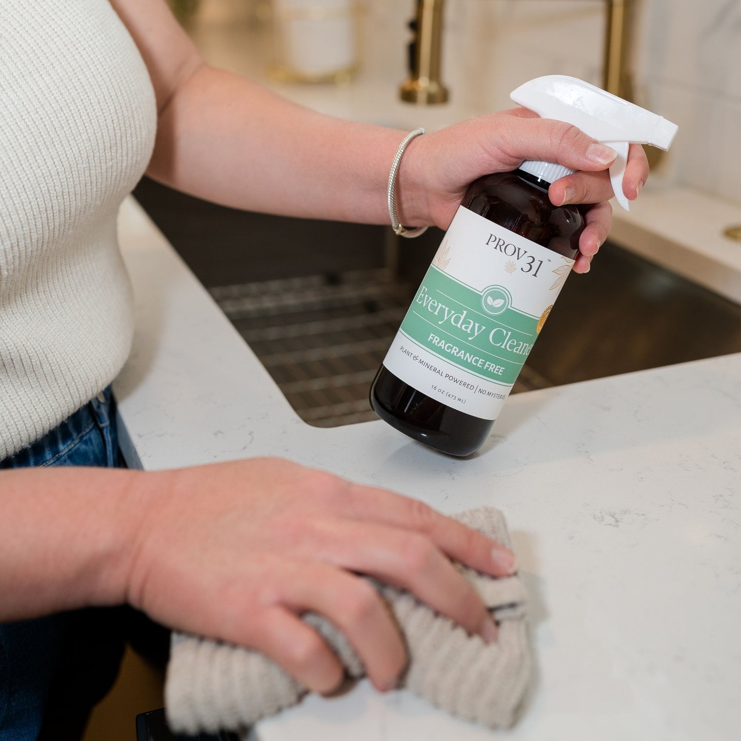 A woman holding a bottle of PROV 31 Everyday Cleaner while wiping a white countertop with a cloth in a kitchen setting.