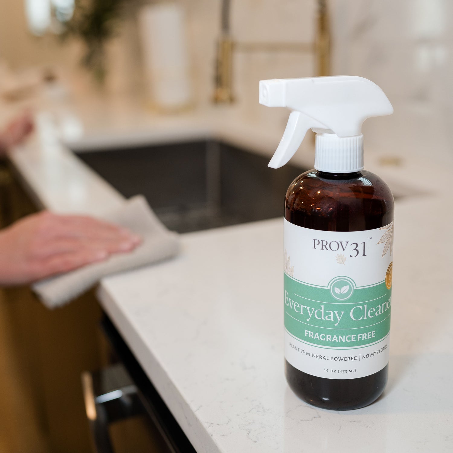 A bottle of PROV 31 Everyday Cleaner spray with a white trigger sprayer and amber bottle, placed on a white kitchen countertop with a blurred kitchen background and a hand wiping down the counter in the distance.