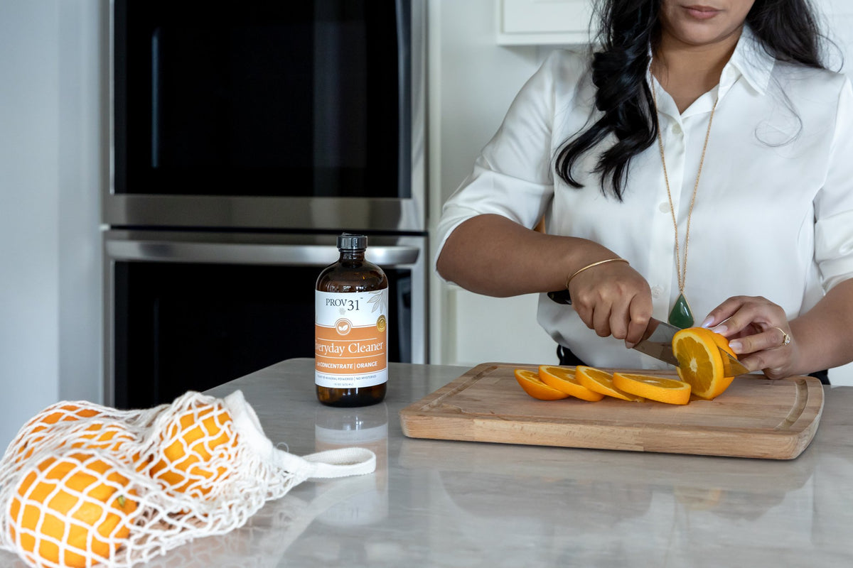 A woman in a white shirt slices an orange on a wooden cutting board in a modern kitchen. A bottle of PROV 31 Everyday Cleaner in Orange, and a white mesh bag of oranges, sit on the counter nearby.