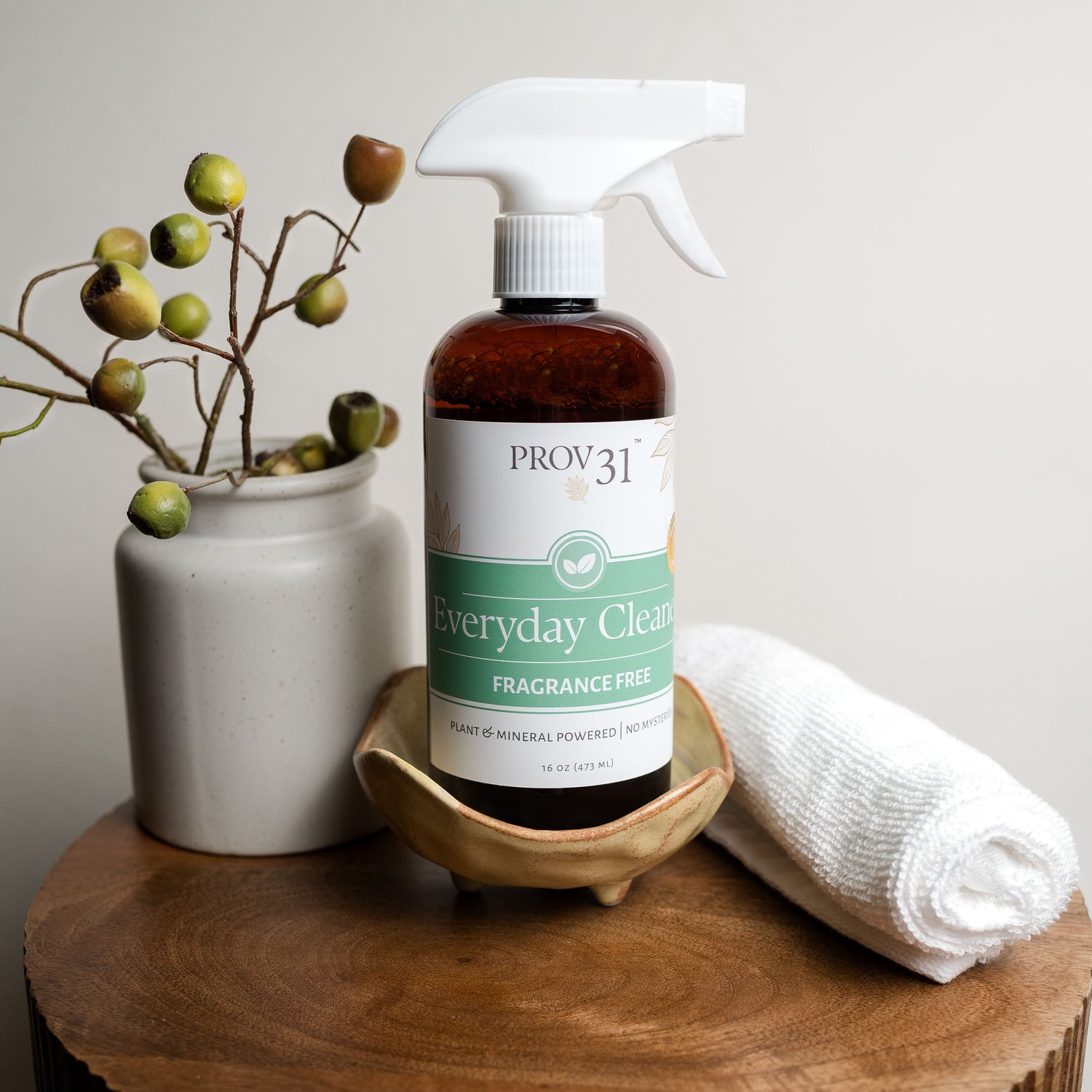A bottle of PROV 31 Everyday Cleaner fragrance-free spray with a white trigger sprayer and amber glass bottle, styled on a wooden table with a white ceramic vase of greenery and a rolled white towel.