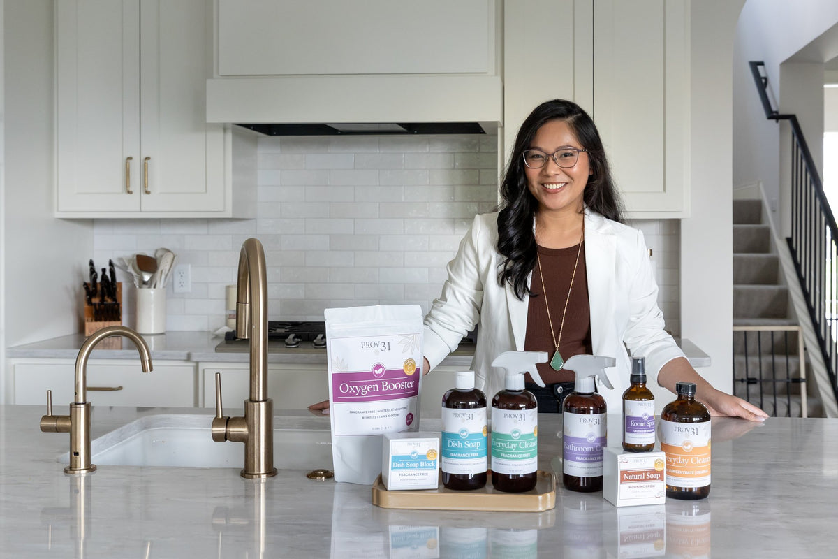 PROV 31's founder stands in a bright kitchen behind a countertop displaying a variety of household products from the brand: spray cleaners, laundry booster, and soaps.