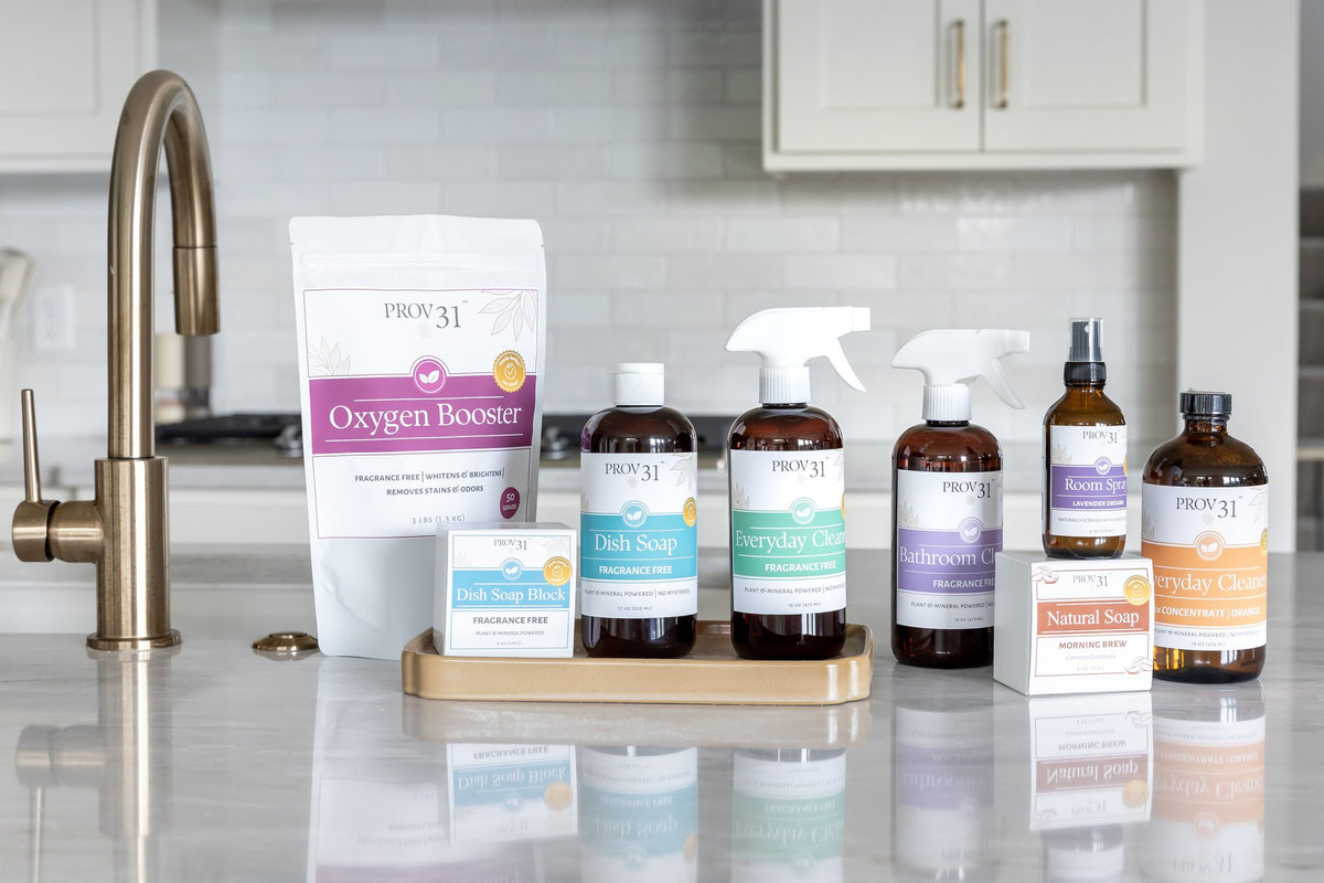 A collection of PROV 31 cleaning products arranged on a gold tray and countertop in a bright kitchen setting. The products include Oxygen Booster, Everyday Cleaner, Bathroom Cleaner, Natural Soap, Room Spray, and Dish Soap.