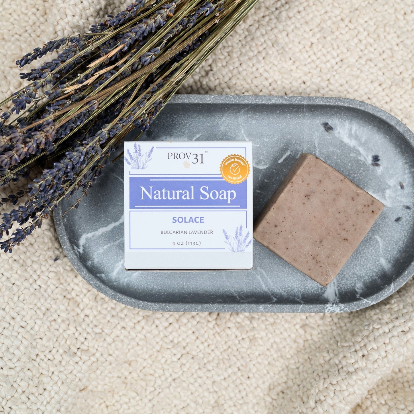 A bar of natural soap from the brand PROV31, labeled 'Solace', rests on a gray stone tray next to dried lavender stems on a textured beige fabric background.