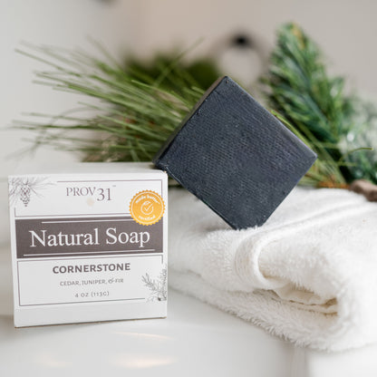 A bar of dark charcoal soap leans against its white and gray packaging box, which reads 'PROV 31 Natural Soap Cornerstone', set on a white surface next to a white towel and green pine branches.