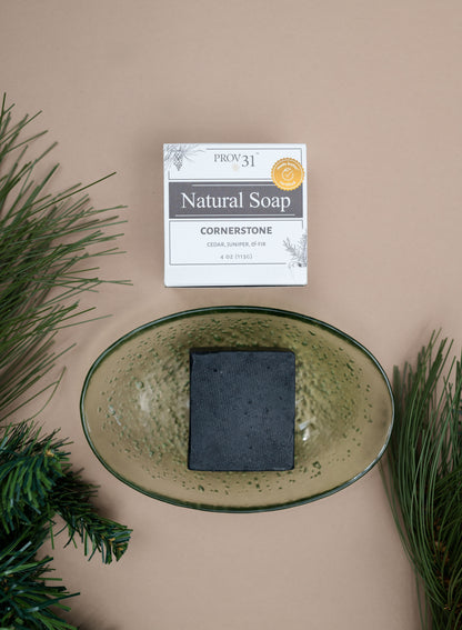 A bar of PROV 31 soap in a speckled green bowl with branded box packaging above, styled with pine branches on a beige background. The box reads 'PROV 31 Natural Soap Cornerstone'.
