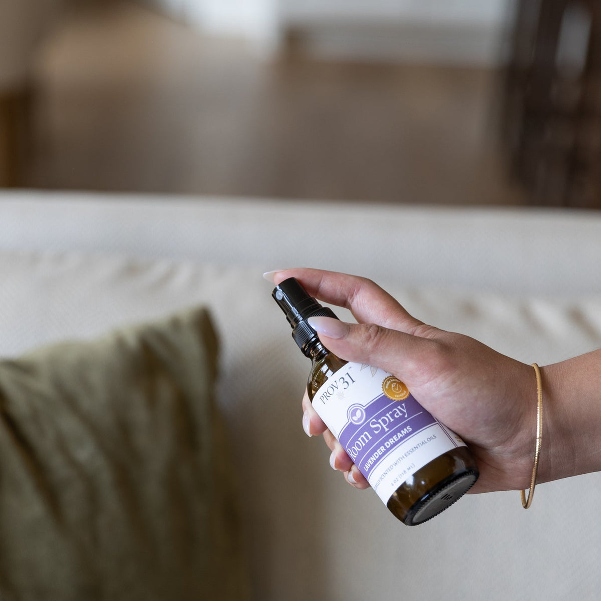 A hand holding a glass amber spray bottle labeled "Room Spray: Lavender Dreams" in front of a beige sofa with a green pillow.