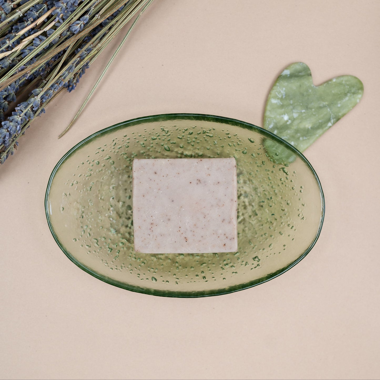 A textured, speckled olive green oval dish holding a square bar of soap, styled with dried lavender and a green gua sha on a beige background.