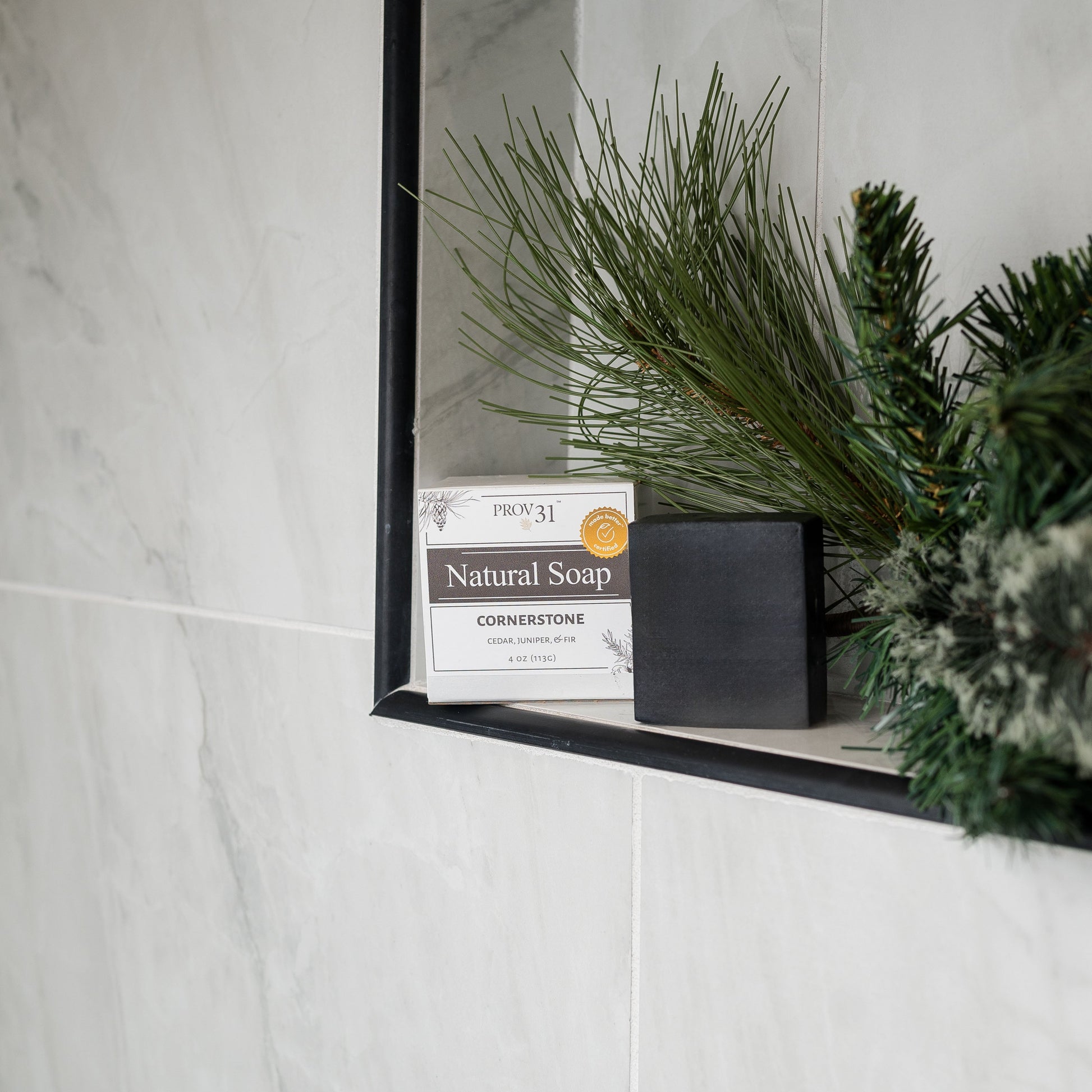 PROV 31 Natural Soap made with charcoal, alongside green pine branches and set inside a light marble-tiled shower wall.