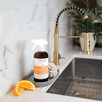 A bottle of PROV 31 Everyday Cleaner Orange scent sits on a marble countertop next to orange slices in a modern kitchen setting with a gold faucet and white subway tiles.