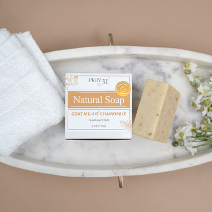 A bar of PROV 31 natural soap and its packaging box, styled on a white marble tray with a folded white towel and chamomile flowers against a solid beige background.